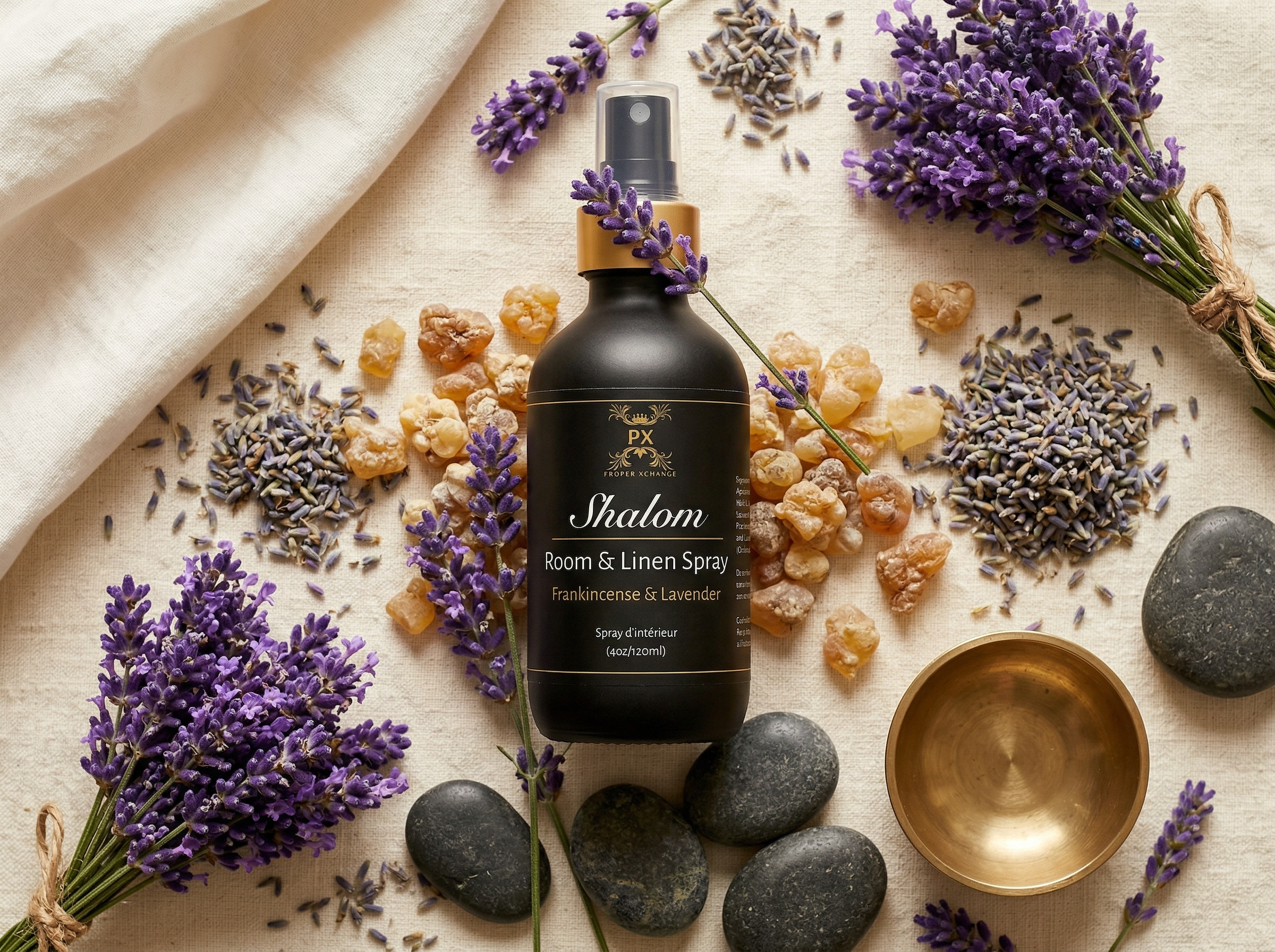 Shalom room & linen spray bottle surrounded by lavender flowers, stones, and a bowl on a light fabric background.