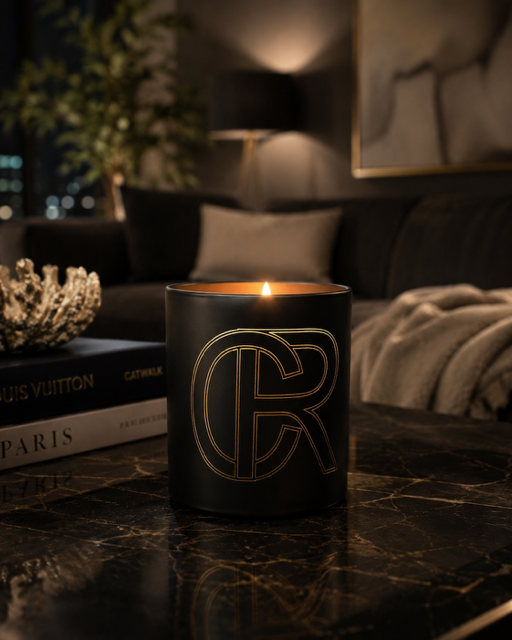 A black candle jar with a gold outlined monogram logo sits lit on a dark marble table in a cozy, dimly lit living room setting.