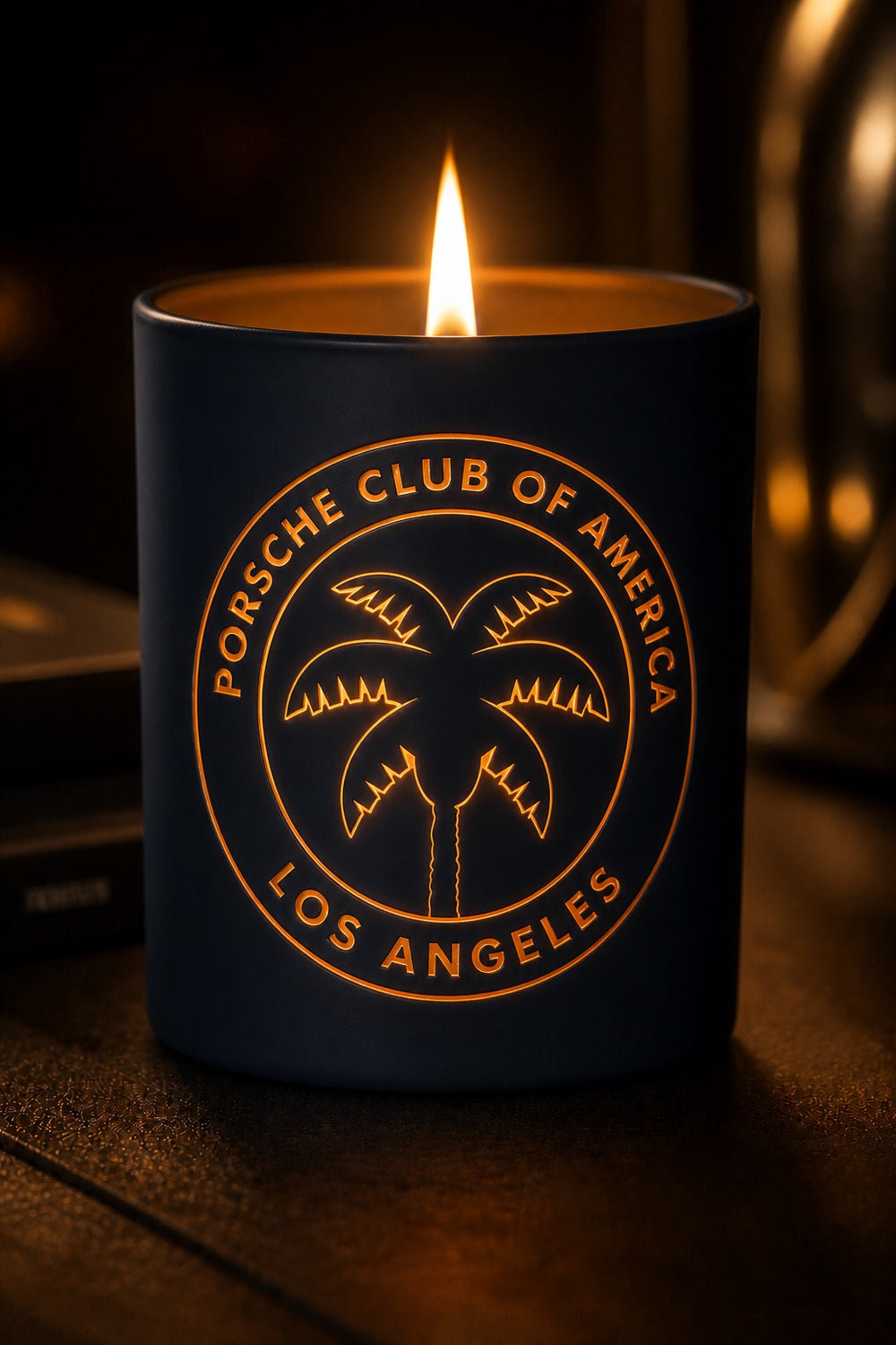 A dark navy blue candle with a lit flame, featuring a gold circular 'Porsche Club of America Los Angeles' palm tree logo on the front, set against a dark, warm-toned background.